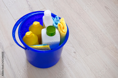 Detergents in plastic bottles, sponges, brush in blue household bucket. Hand in yellow rubber glove. Service concept. Copy space. © Ga_Na
