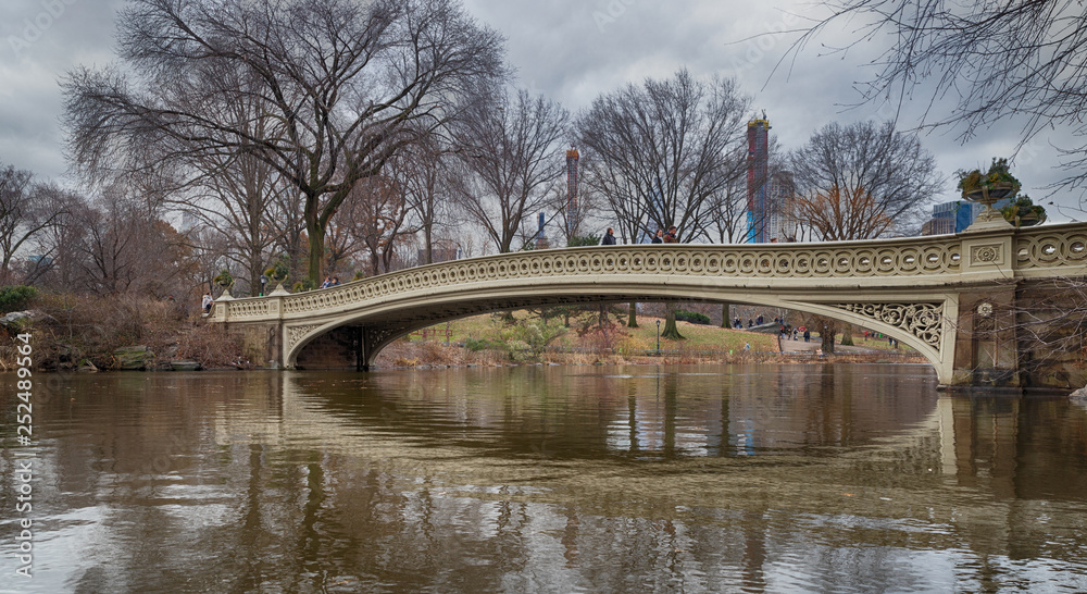 The bow bridge in central park, New York city daylight view with reflection in water and clouds,trees and Manhattan buildings in background