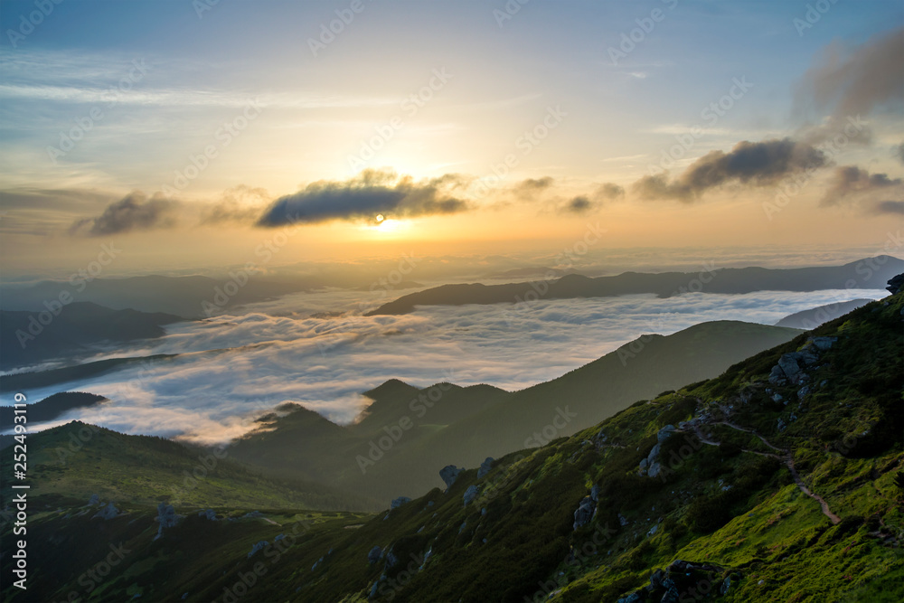 Fantastic view of mountain valley covered with low white puffy like snow clouds stretching to foggy horizon under bright morning sky with light orange glow at sunrise. Beauty of nature concept.