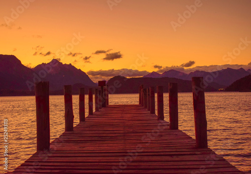viejo muelle sobre un lago rodeado de montañas sobre el cual se reflejan los ultimos rayos de sol
