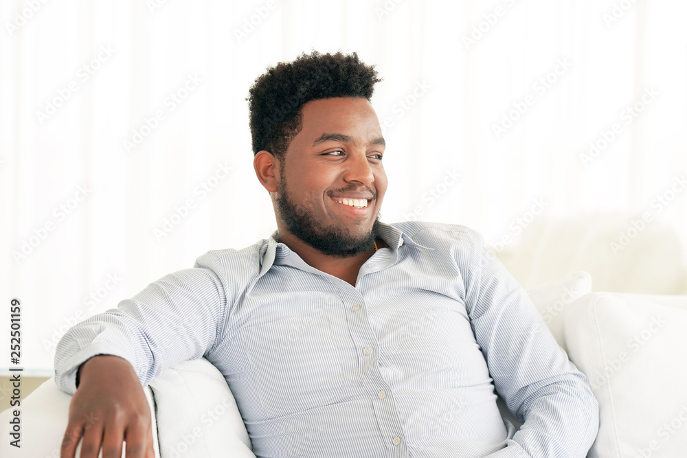Good looking young African guy sitting on sofa and smiling. Lifestyle Asian man at home. Man Relaxing On Sofa At Home. Good looking young African guy sitting on sofa and smiling. Lifestyle Asian man at home. Man Relaxing On Sofa At Home.