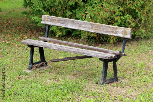 Broken old public bench with dilapidated wooden boards and strong metal supports surrounded with freshly cut grass and dense bush in background on warm sunny day
