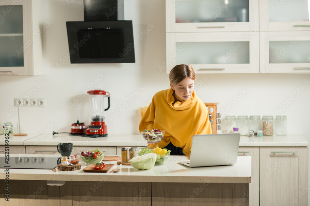 Attentive woman using laptop in the kitchen while standing with salad Attentive woman using laptop in the kitchen while standing with salad