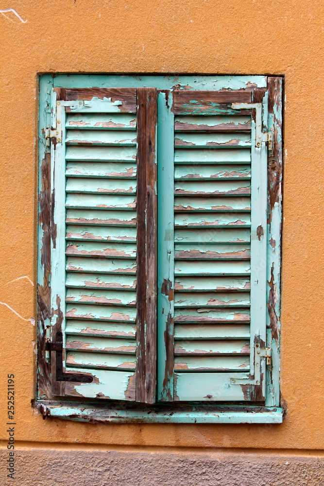 Dilapidated old closed wooden blinds with cracked blue paint mounted with rusted metal hinges on wooden frame surrounded with orange and purple facade of family house wall on warm sunny day