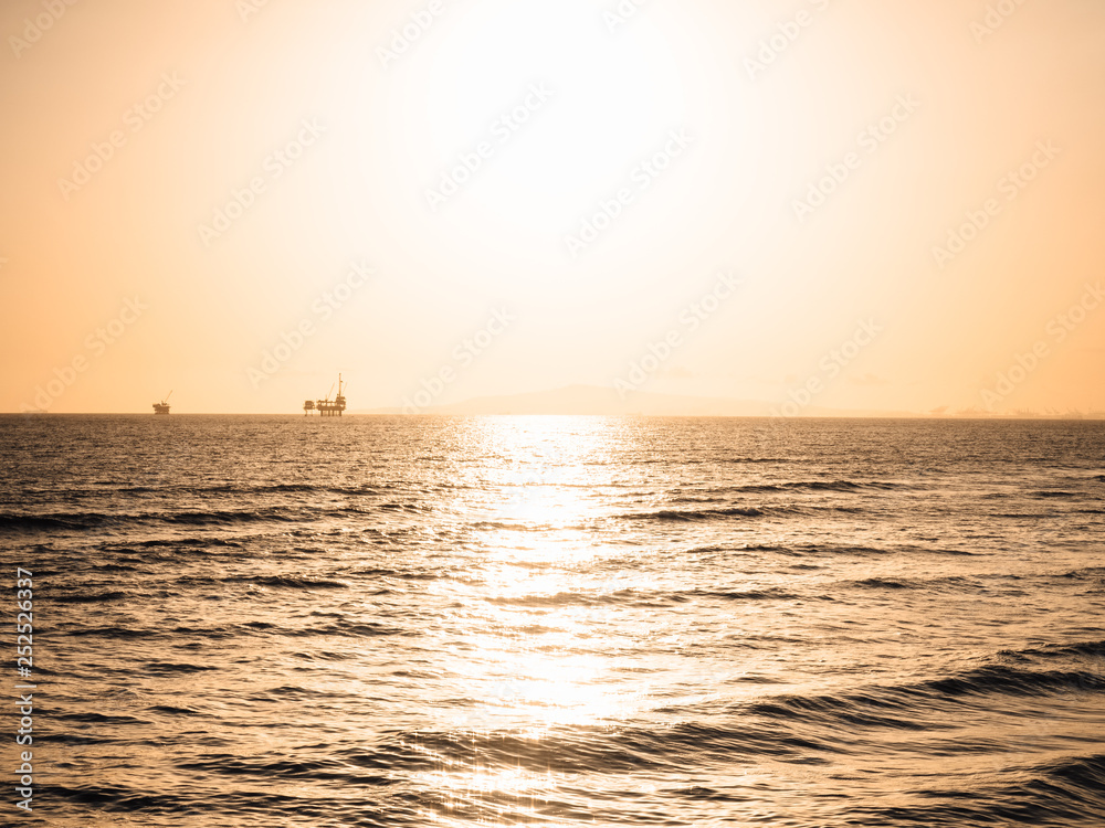 Pacific Ocean during sunset at Huntington Beach, Orange Country, California, USA with view to offshore oil rig platforms drilling for petroleum and gas, container ships and golden sky in background