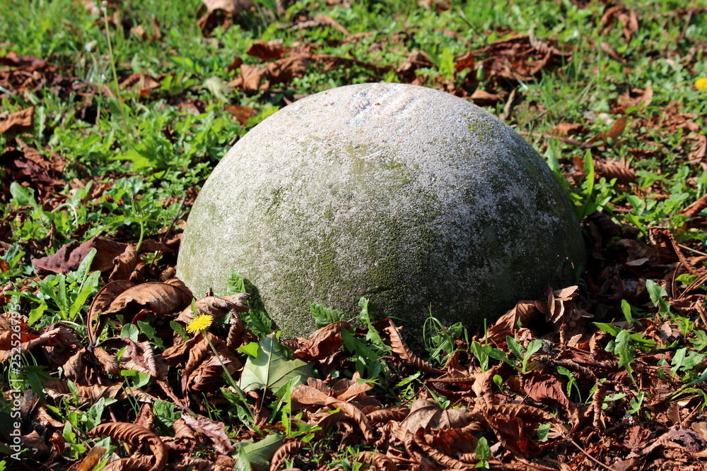 Half of grey concrete sphere barrier surrounded with green grass and fallen brown leaves preventing cars from entering local park on warm sunny day Half of grey concrete sphere barrier surrounded with green grass and fallen brown leaves preventing cars from entering local park on warm sunny day