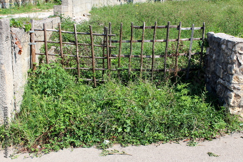 Homemade improvised backyard doors made from wooden sticks mounted on traditional stone wall blocking entrance to overgrown backyard of abandoned house on warm sunny day