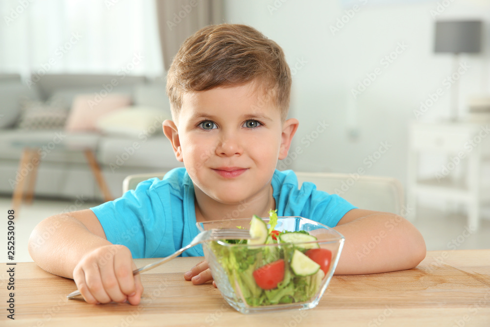 Adorable little boy eating vegetable salad at table in room Adorable little boy eating vegetable salad at table in room