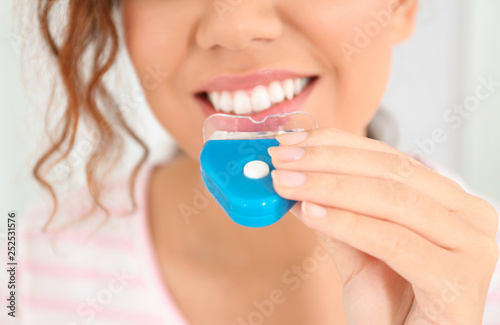 Young African-American woman using teeth whitening device on light background, closeup