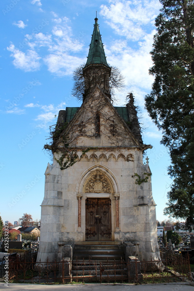 Large highly decorated carved stone family tomb with strong wooden doors locked with metal padlock surrounded with rusted metal fence next to tall tree on cloudy blue sky background