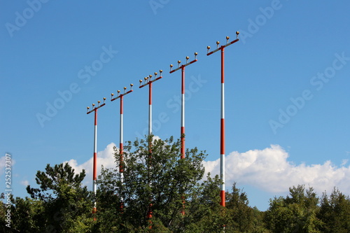 Multiple high metal red and white poles with airport runway guiding lights surrounded with dense trees on cloudy blue sky background