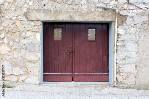 Old wooden garage doors made of wooden boards and rubber rain protection locked with rusted latch and padlock with two small windows mounted on traditional stone wall next to water pipe on warm cloudy