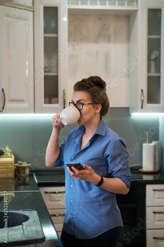 Young attractive girl drinks coffee in kitchen. She is holding cell phone. Good morning.