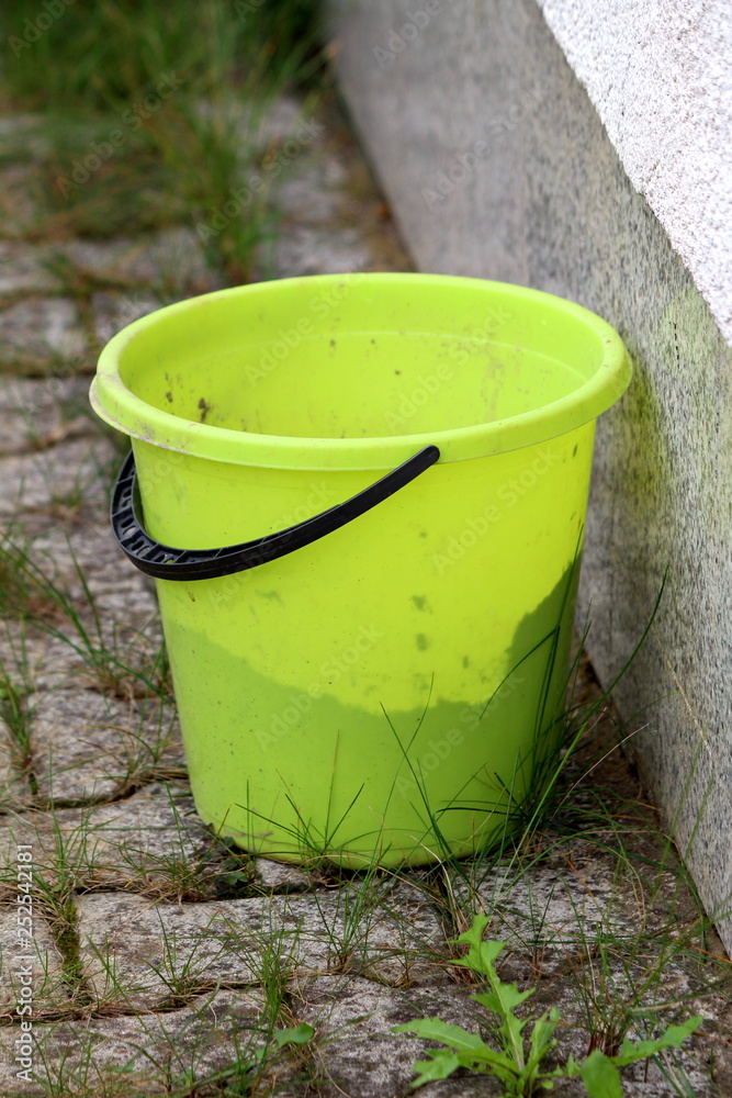 Plastic bucket with black handle left behind house on stone tiles with grass growing between on warm sunny day Plastic bucket with black handle left behind house on stone tiles with grass growing between on warm sunny day