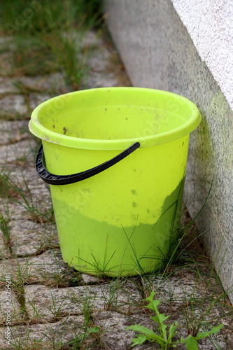 Plastic bucket with black handle left behind house on stone tiles with grass growing between on warm sunny day