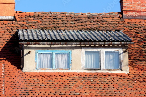 Roof windows with light blue and white dilapidated wooden frames surrounded with small roof tiles and rusted gutter next to two red brick chimneys on clear blue sky background