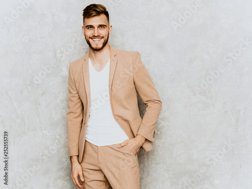 Portrait of handsome smiling hipster lumbersexual businessman model wearing casual beige suit. Fashion stylish man posing against gray wall photo