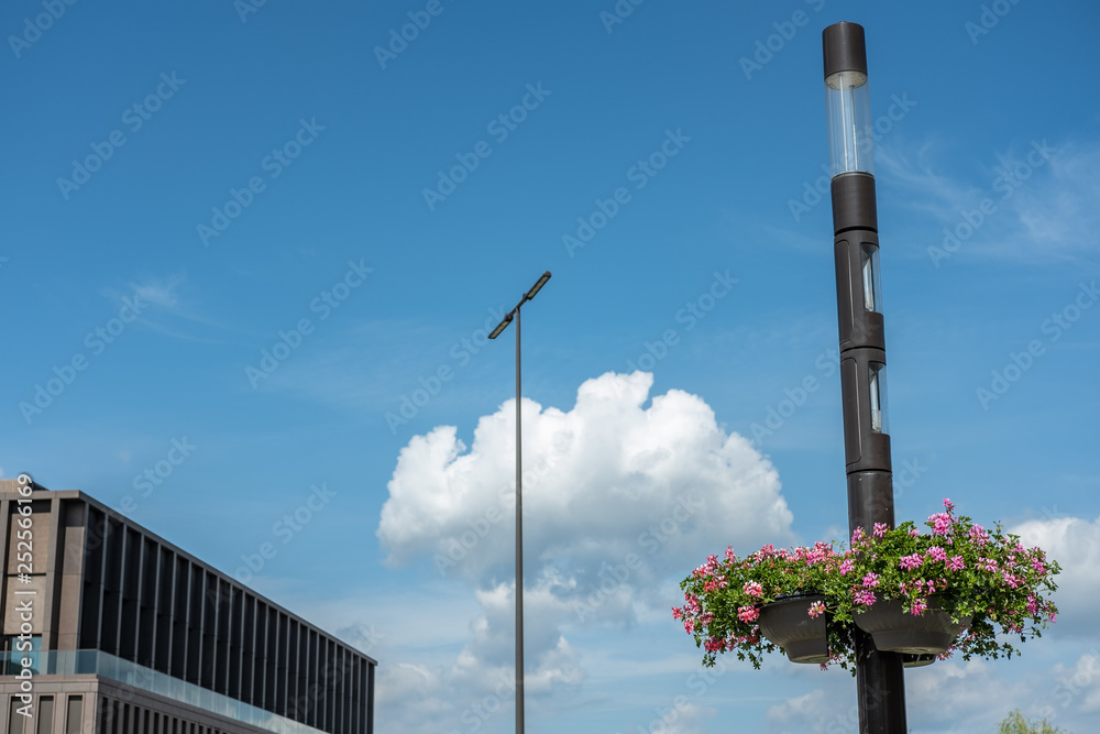 Moderne Strassenlaterne mit Blumenschmuck vor Stadthalle mit blauem Himmel und Wolken Moderne Strassenlaterne mit Blumenschmuck vor Stadthalle mit blauem Himmel und Wolken