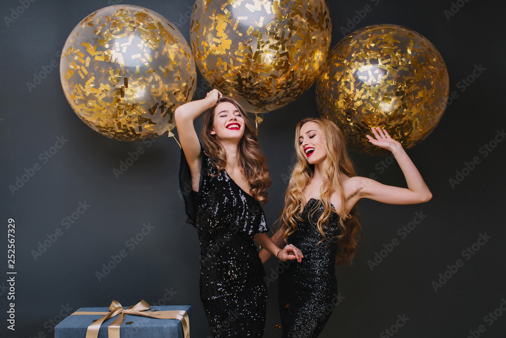 Naklejka premium Beautiful ladies dancing with hands up in front of shining helium balloons and smiling. Indoor photo of refined brown-haired birthday girl chilling with friend and laughing.