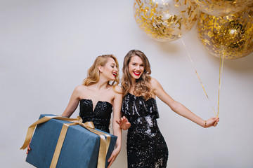 Attractive ypung women in black luxury dresses celebrating birthday party with big present and balloons on white background. Excited, having fun, charming models, celebrating, smiling