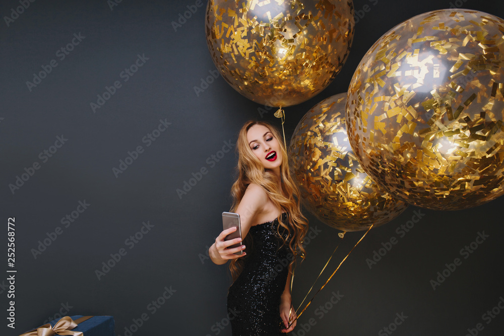 Naklejka premium Indoor portrait of stunning fair-haired girl making selfie at festive. Spectacular woman with party balloons having fun and taking photo of herself on black background.