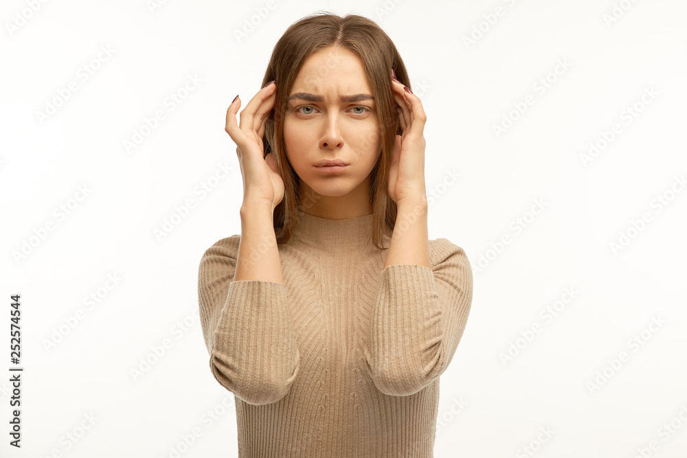 Upset woman looks at camera, keeps hands on temples, raises eyebrow, has long dark hair, dressed in light sweater, model against white wall Upset woman looks at camera, keeps hands on temples, raises eyebrow, has long dark hair, dressed in light sweater, model against white wall