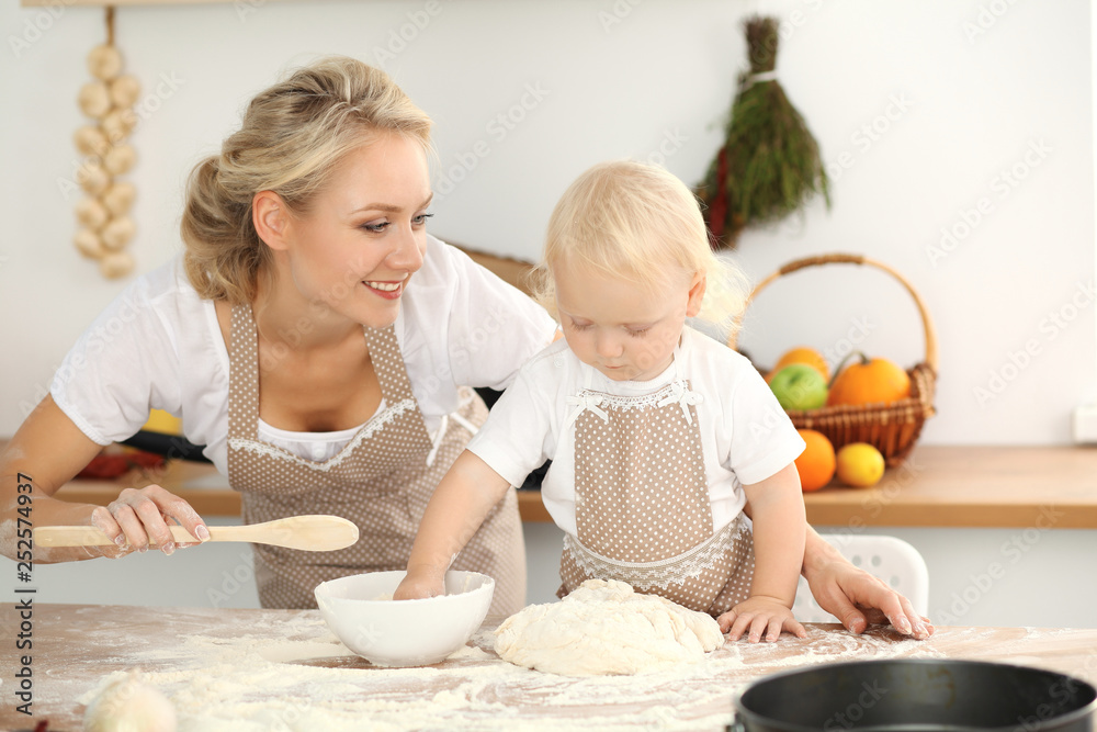 Little girl and her blonde mom in beige aprons playing and laughing while kneading the dough in kitchen. Homemade pastry for bread, pizza or bake cookies. Family fun and cooking concept Little girl and her blonde mom in beige aprons playing and laughing while kneading the dough in kitchen. Homemade pastry for bread, pizza or bake cookies. Family fun and cooking concept