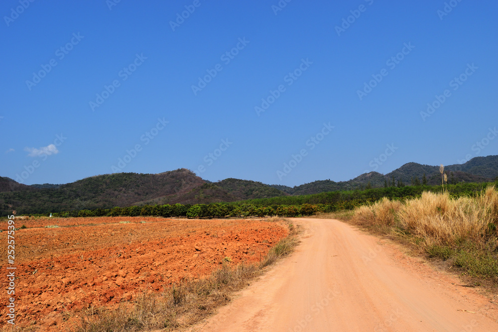 Open dirt road in farmland with mountain forest and blue sky in background, The land was plowing and turning over the soil in preparation for planting, Thailand