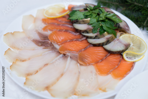 Fresh slices of salmon, red fish and herring on a white plate in a restaurant. Fish platter is decorated with slices of fresh lemon, herbs.