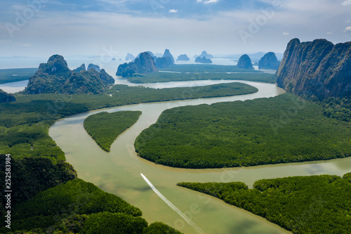 Aerial drone view of a small boat on a mangrove forest river heading towards open ocean past towering limestone cliffs and islands