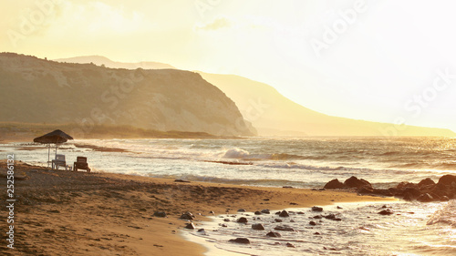 Two sunbeds on empty wild beach lit by golden sunset. Small waves over rock  hills in hazy background