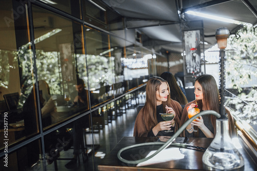 Young beautiful girls are sitting on the summer terrace of a modern cafe and drinking refreshing cocktails in orange and green. © Тарас Нагирняк