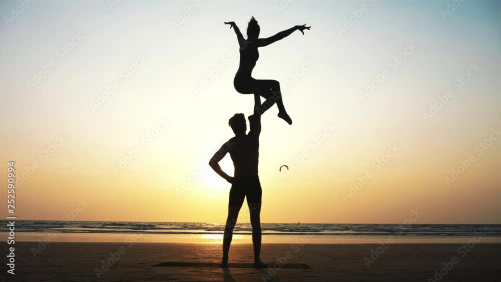 Silhouette of fit sporty couple practicing acrobatic yoga with partner together on the beach. Male standing and holding his female partner over his head. Mix race couple doing acrobatic exercise. Silhouette of fit sporty couple practicing acrobatic yoga with partner together on the beach. Male standing and holding his female partner over his head. Mix race couple doing acrobatic exercise.