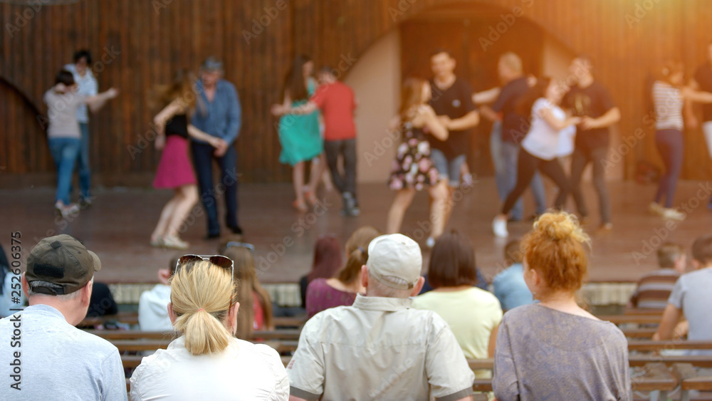 Group of mature people at outdoor concert. Couple sitting outdoors and looking at performance on the stage, back view. Holidays, leisure and lifestyle.