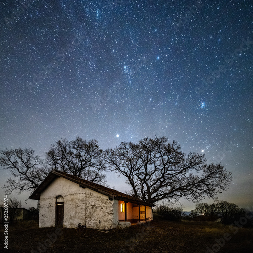 A small old church with a lighted window at night. Behind the church is an old tree without leaves. Starry Sky with Milky Way. © Mincho Minchev