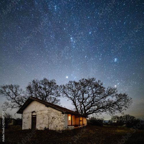 A small old church with a lighted window at night. Behind the church is an old tree without leaves. Starry Sky with Milky Way.