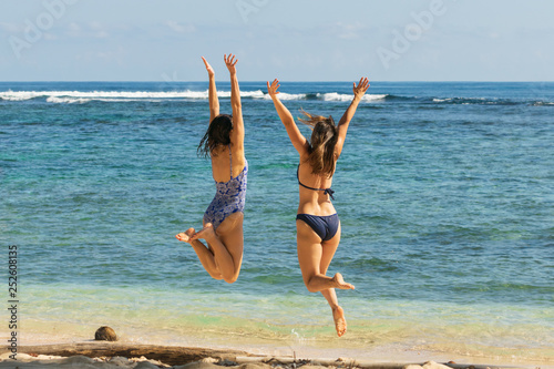 Two attractive joyful girls in blue bikini and long hair jumping on the beach with hands outstretched expressing happiness in vacation.
