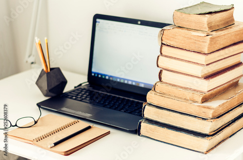 Laptop, stack of old books, notebook and pencils on white table, education retro concept background.