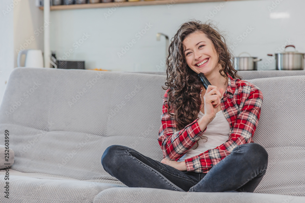 A beautiful woman watches the TV and is sitting on the couch and holds the remote control in his hand. A brunette in a shirt looks at the TV with long hair. A beautiful woman watches the TV and is sitting on the couch and holds the remote control in his hand. A brunette in a shirt looks at the TV with long hair.