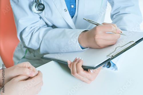 Female doctor arm hold silver pen filling patient complaints list clipped to pad. Female patient tells the doctor about her problems. Physical exam or disease prevention concept.