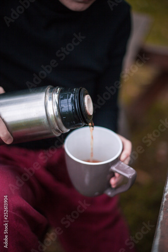 man holding a сup of tea and a thermos on a picnic in the woods