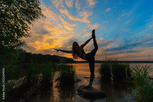 Blonde woman doing bow pulling pose outdoors on a stone in the lake. Yoga nature concept.