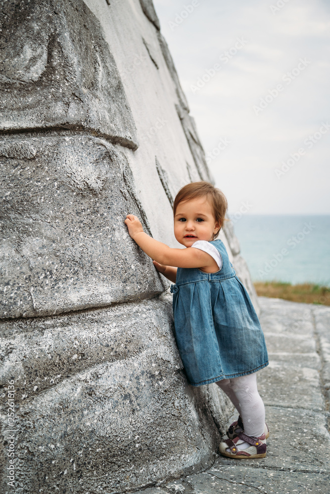 Happy small baby girl in jeans dress standing near to the wall of the old lighthouse Happy small baby girl in jeans dress standing near to the wall of the old lighthouse