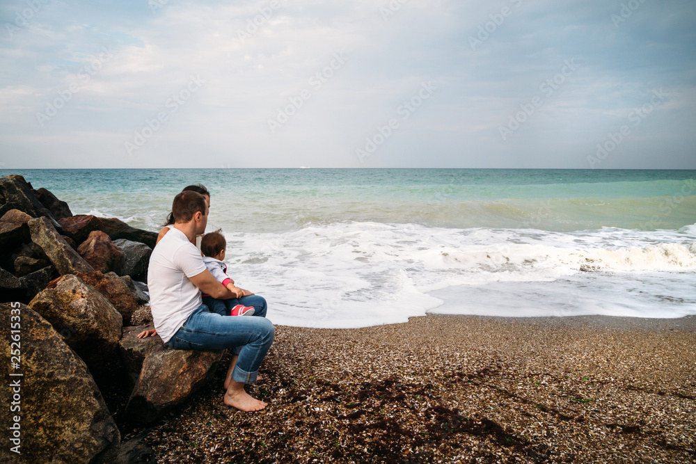 Happy young family in white t-shirts and blue jeans with a small daughter in blue dress sitting at the seaside Happy young family in white t-shirts and blue jeans with a small daughter in blue dress sitting at the seaside