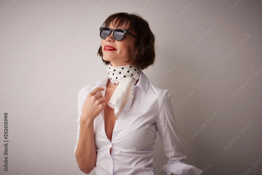 Fashionable young woman studio portrait. Attractive woman wearing sunglasses and scharf while posing at light grey background. Fashionable young woman studio portrait. Attractive woman wearing sunglasses and scharf while posing at light grey background.