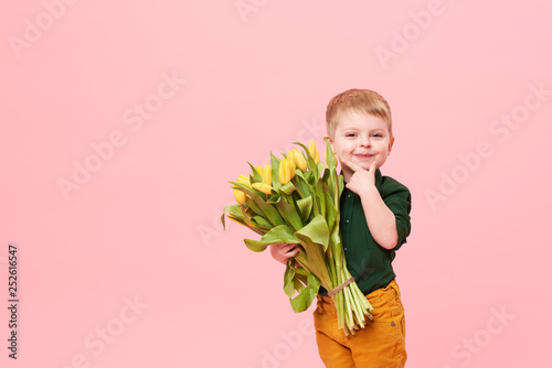 Adorable smiling child with spring flower bouquet looking at camera isolated on pink. Little toddler boy holding yellow tulips as gift for mom. Copy space for text on left side