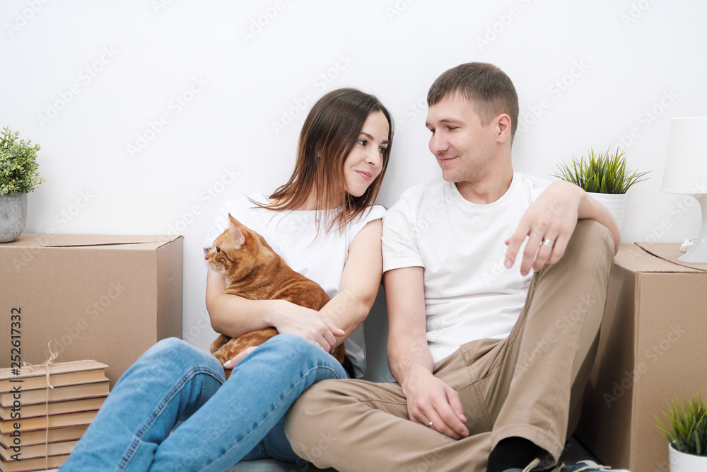 A young married couple with a red cat, a man and a woman, are sitting on the floor in a bright room against the background of cardboard boxes, green plants and things.