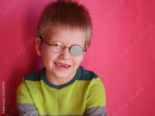 Fototapeta Naklejka Na Ścianę i Meble -  Portrait of funny child in new glasses with patch for correcting squint on pink background. boy with lost baby teeth..Ortopad Boys Eye Patches nozzle for glasses for treatment of strabismus (lazy eye)
