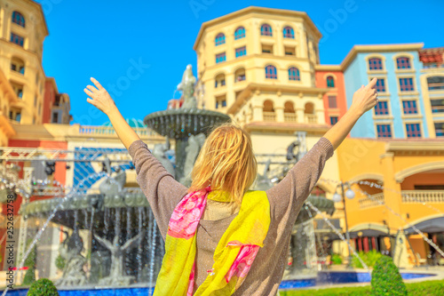 Carefree blonde woman enjoys water fountains in Central Medina, an exclusive residential area in The Pearl Qatar, icon of Doha, Persian Gulf, Middle East, Arabian Peninsula. Souq Medina on background.