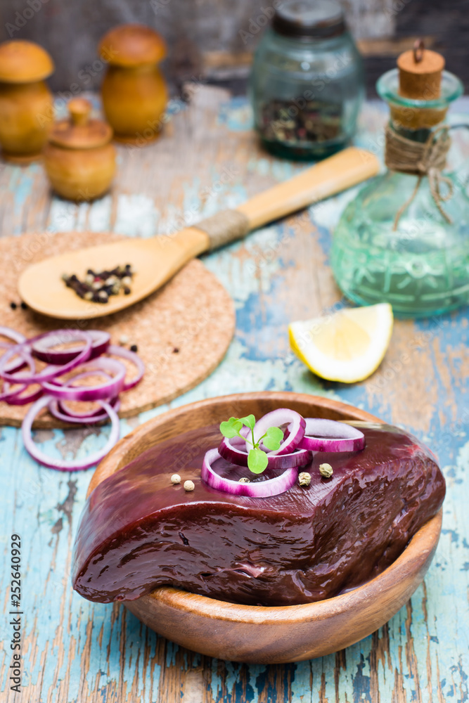 A piece of raw beef liver in a bowl and spices for cooking on a wooden table